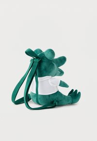 Πράσινο σακίδιο πλάτης σε σχήμα κροκοδείλου από μαλακό ύφασμα με ρυθμιζόμενους ιμάντες και λευκό πουκάμισο, εμφανίζεται καθιστό σε ορθή θέση πάνω σε απλό φόντο.