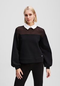 Sweatshirt noir avec une partie supérieure transparente et un col blanc. Présente des manches longues et bouffantes ainsi qu'un ourlet ajusté. Détail brodé minimaliste.