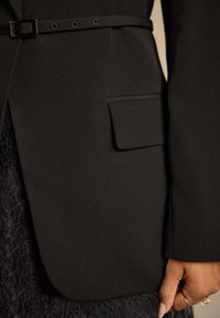 Black tailored blazer featuring a slim fit, a belted waist, and a single front pocket. The fabric has a smooth finish with no visible patterns.