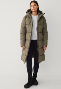 Frau in einem olivgrünen, langen gepolsterten Mantel, hellgrauem Sweatshirt, schwarzen Leggings und schwarzen Stiefeletten, die vor einem weißen Hintergrund steht.