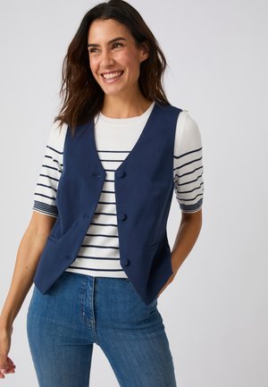 Femme souriante portant un gilet bleu marine boutonné sur une chemise blanche à rayures horizontales bleu marine et un jean bleu, debout devant un fond uni.