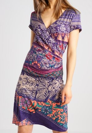 Kobieta ubrana w krótką sukienkę z krótkim rękawem i dekoltem w serek, o długości do kolan, z kolorowym kwiatowym i geometrycznym wzorem w odcieniach fioletu, pomarańczy, różu i zieleni.