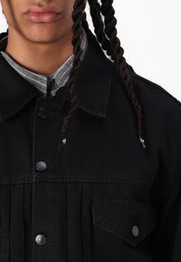 Svart denimjakke med knappestolpe foran, spiss krage og brystlommer; har teksturert stoff og sølvfargede detaljer.