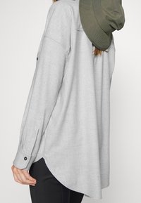 Camisa de franela gris con dobladillo redondeado, puños de botón y aberturas laterales; combinada con un sombrero verde colgado en la parte de atrás.