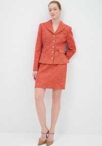 Roter Tweed-Anzug mit tailliertem Blazer mit Schalkragen, goldenen Knöpfen und einem Bleistiftrock. Glatte Textur mit dezentem Muster.