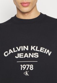 Felpa navy con colletto a costine che mostra "CALVIN KLEIN JEANS" in bianco, 1978, e logo cK. Texture morbida, design casual.