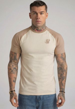 Mann med tatoveringer på nakke og armer iført beige raglan-skjorte og blå jeans, stående foran ensfarget bakgrunn.