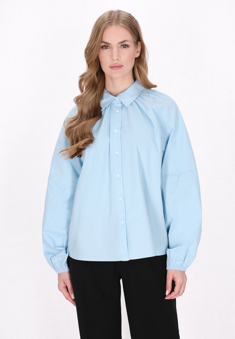 Camicia azzurro chiaro a maniche lunghe con colletto, dettagli arricciati sulle spalle e polsini elasticizzati. Pantaloni neri sotto.