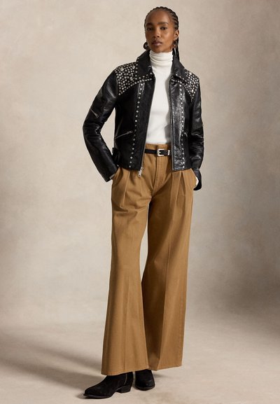 Femme debout, les mains dans les poches, portant une veste en cuir noire cloutée, un col roulé blanc, un pantalon large beige et des bottines noires.