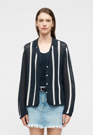 Vrouw draagt een marineblauwe opengewerkte cardigan met witte verticale strepen over een zwarte top en een lichtblauwe denimrok met rafels.