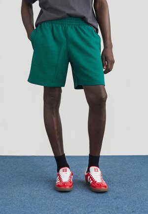 Zelené bavlnené šortky s elastickým pásom a bočnými vreckami. Nosia sa s červeno-bielymi teniskami na modrom povrchu.