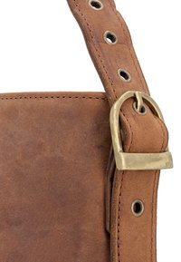 Bolso de cuero de ante marrón con correa ajustable sujeta por una hebilla de latón y ojales de metal sobre un fondo blanco.