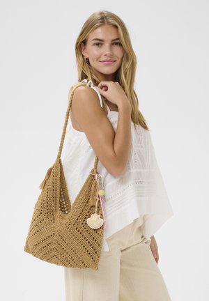 Femme blonde portant un haut blanc sans manches et un pantalon beige, avec un grand sac à bandoulière en crochet marron orné d'un porte-clés coquillage.