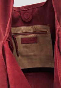 Die Innenseite der roten Wildleder-Tasche verfügt über eine hellbraune Stoffauskleidung, eine Reißverschlusstasche und ein geprägtes Lederschild mit glatter Textur.