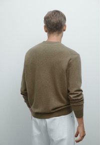 Maglione di lana marrone con collo a giro, maniche aderenti e orlo a coste, abbinato a pantaloni chiari, visto da dietro.