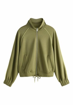 Kobieta z długimi warkoczami, ubrana w oliwkową kurtkę na zamek i dopasowane spodnie ze sznurkiem, stoi z rękami w kieszeniach na szarym tle.