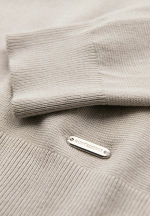 Pull en tricot beige avec une texture côtelée et une étiquette argentée gravée de "QUINTESSENCE". Gros plan sur le détail du poignet replié.