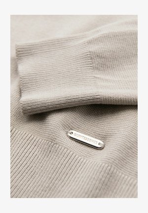 Pull en tricot beige avec une texture côtelée et une étiquette argentée gravée de "QUINTESSENCE". Gros plan sur le détail du poignet replié.
