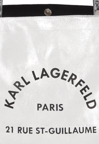 Sac fourre-tout blanc brillant avec texte noir. Le logo imprimé indique "KARL LAGERFELD PARIS" avec l'adresse en dessous. Fermeture magnétique en haut.