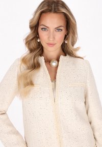 Tweedjacke in Creme mit metallischen Fadenakzenten, mit einer strukturierten Oberfläche und einem Reißverschluss. Getragen mit einer großen Perlenkette.