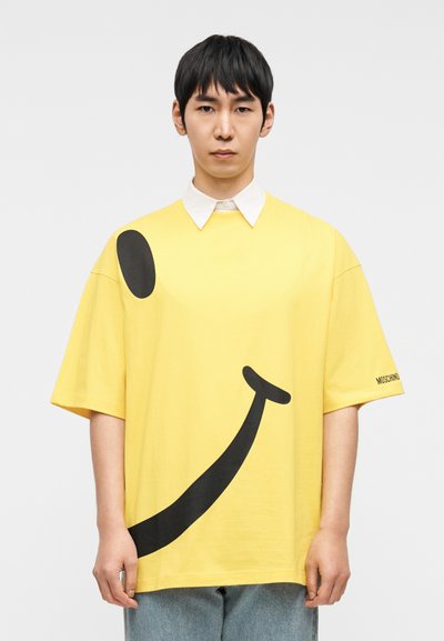 Κίτρινο υπερμεγέθες μπλουζάκι με μεγάλο μαύρο γραφικό χαμογελαστού προσώπου. Διαθέτει λευκή λαιμόκοψη και επωνυμία "MOSCHINO" στο μανίκι. Καθημερινό στυλ.