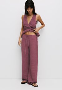Dvoudílný outfit z texturované mauve látky; bezramenný zavinovací top s hlubokým výstřihem do V a odpovídající široké kalhoty s elastickým pasem a šňůrkou.