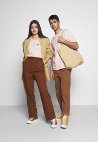 Två modeller bär avslappnade kläder: en ljusrosa t-shirt, en beige rutigt blazer, bruna högmidjade byxor och färgglada sneakers. En av dem håller en beige tote-väska.