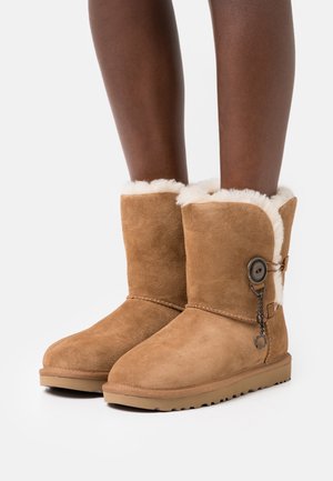 Bottes mi-mollet en daim marron clair, doublées de polaire blanche, avec un grand bouton et un détail de charme, portées sur des jambes nues sur un fond blanc uni.