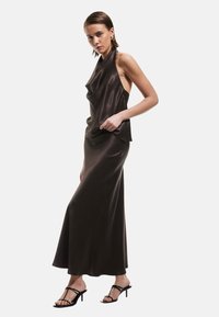 Abito lungo in raso marrone con scollo all'americana, design drappeggiato e vestibilità slim. Il modello indossa tacchi neri con cinturini, mettendo in risalto la silhouette dell'outfit.