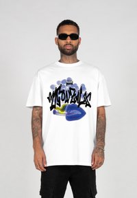Hvid bomulds T-shirt med fed sort graffiti-lignende tekst og en blå grafisk illustration. Modellen bærer sorte shorts og solbriller.