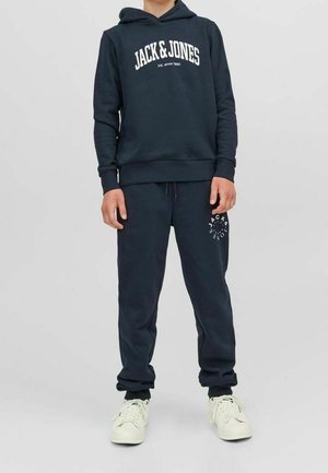 Laivastonsininen huppari, jossa valkoinen "JACK & JONES" -teksti, yhdistettyyn vastaaviin jogger-housuihin, joissa on logoyksityiskohta. Kengät ovat valkoiset ja nauhalliset.