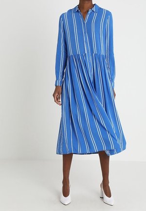 Frau trägt ein blaues Midikleid mit weißen vertikalen Streifen, langen Ärmeln und geknöpftem Vorderteil, kombiniert mit weißen High-Heel-Schuhen.