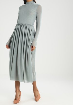 Kobieta ubrana w długą sukienkę midi z długim rękawem, wysokim kołnierzem i jasnoszarym kolorem oraz marszczoną spódnicą, do tego jasnoróżowe buty na wysokim obcasie.