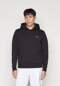 Černá mikina s kapucí vyrobená z měkké tkaniny, s přední kapsou a vyšitým logem "Calvin Klein" na hrudi.