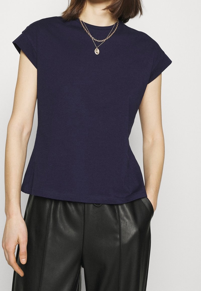 Femme portant un haut à manches courtes bleu marine, un pantalon en cuir noir et des colliers en chaîne dorée superposés avec un petit pendentif.