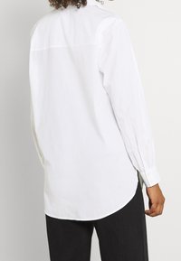 Chemise blanche boutonnée avec manches longues, présentant une coupe décontractée et un ourlet incurvé, fabriquée en coton lisse. Design simple et épuré.