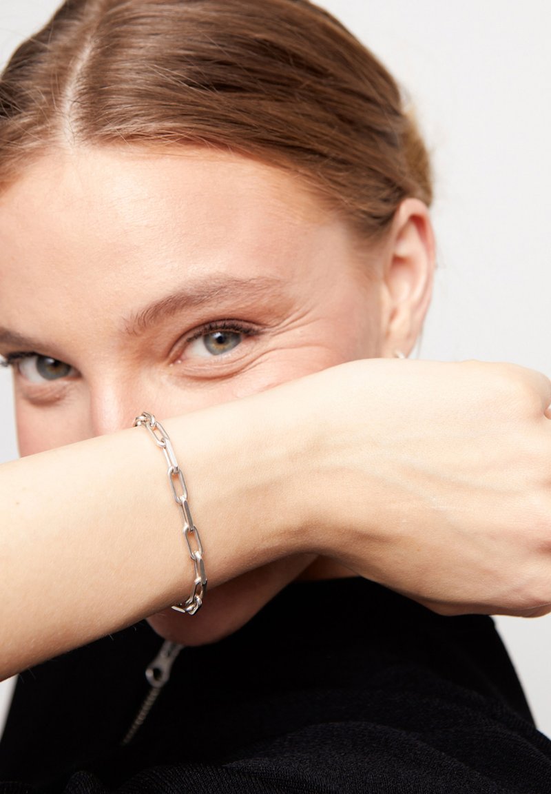 Pulsera de cadena de plata con segmentos alargados entrelazados en una muñeca, mostrando una superficie pulida y un diseño minimalista, sobre un fondo neutro.