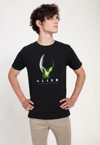 Czarna bawełniana koszulka z grafiką przedstawiającą kosmiczne jajo. Tekst poniżej brzmi „ALIEN” z hasłem w kolorze zielonym. Krótkie rękawy, okrągły dekolt.