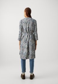 Sukienka z długim rękawem o wzorze paisley w odcieniach niebieskiego i szarości, z wiązaniem w talii, bocznymi rozcięciami i luźnym krojem, zestawiona z jeansami.