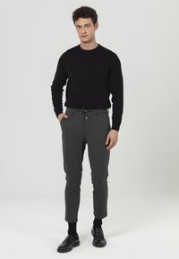Schwarzer Pullover mit langen Ärmeln, kombiniert mit dunkelgrauen, taillierten Hosen. Verfügt über einen elastischen Bund, Taschen und glänzende Knöpfe. Getragen mit schwarzen Slippern.