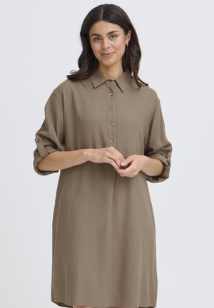 Kobieta o ciemnych włosach, ubrana w luźną, brązową sukienkę z długim rękawem, z podwiniętymi mankietami, stojąca na tle jasnego, jednolitego tła.
