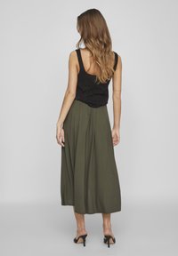 Zwart textuurloze mouwloze top gecombineerd met een lange, vloeiende olijfgroene rok met een losse pasvorm en subtiele plooien. Zwarte hakken.