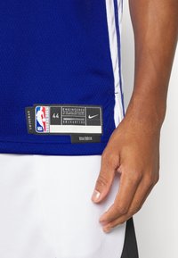 Gros plan d'une personne portant un maillot NBA swingman bleu avec une étiquette Nike et un short blanc.