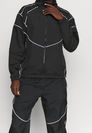 Man draagt een zwarte trainingspak met witte biezen, capuchon op, hand dicht bij mond, staand tegen een effen achtergrond.