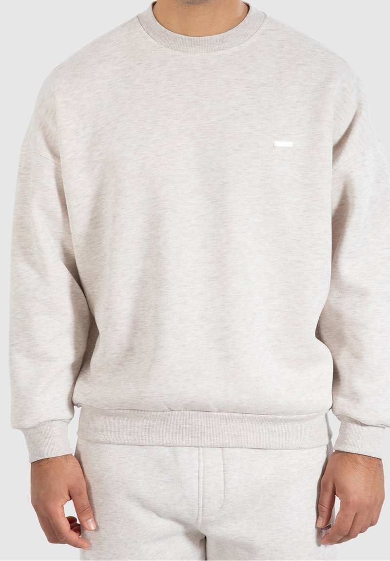 Homme portant un sweat-shirt gris clair à manches longues et un pantalon assorti, présenté des épaules aux hanches sur un fond uni.