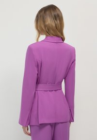 Blazer aderente viola con vita alta e cintura, maniche lunghe e texture liscia. Presenta un colletto classico e un dettaglio plissettato sulla schiena.