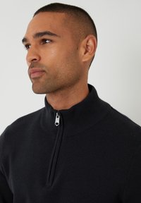 Czarny sweter z dzianiny z wysokim kołnierzem i zamkiem na ćwiartkę, gładką teksturą oraz prążkowanymi akcentami na kołnierzu i dolnym brzegu.