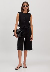 Zwart mouwloos cropped top met knopen, gecombineerd met zwarte culottes. Het model houdt een zwarte clutch vast en draagt smalle zonnebrillen en slippers.