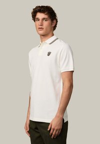 Biała koszulka polo wykonana z bawełny z zapięciem na guziki pod kołnierzykiem, z kontrastowymi ciemnymi wykończeniami i małym haftowanym logo na piersi.