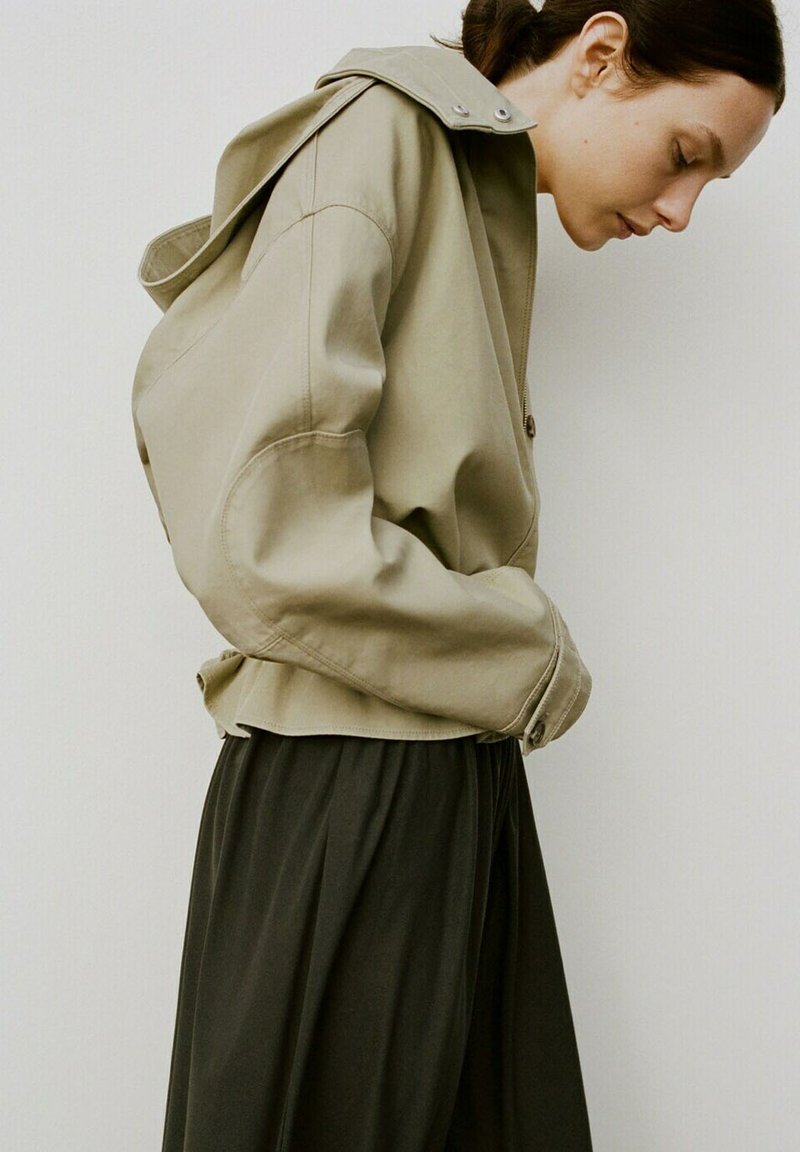 Femme portant une veste beige oversize à capuche et une jupe noire, regardant vers le bas avec les mains dans les poches de la veste sur un fond uni.